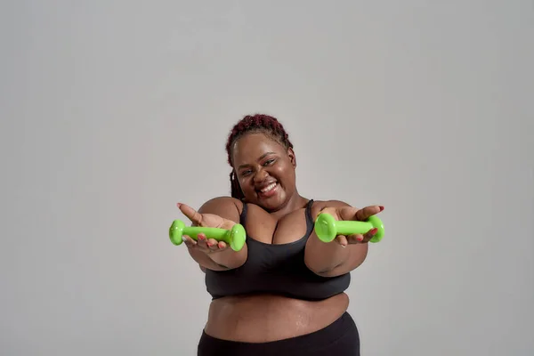 Plump, plus size afrikanisch-amerikanische Frau in Sportbekleidung, die die Arme ausstreckt und grüne Hanteln im Studio vor grauem Hintergrund hält — Stockfoto
