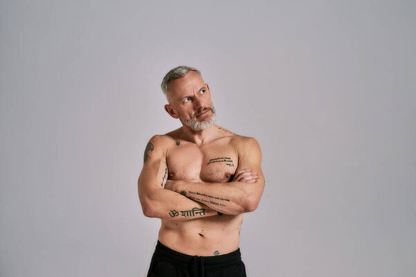 Half naked middle aged muscular man looking aside, showing his body, while posing in studio over grey background. Front view. Horizontal shot