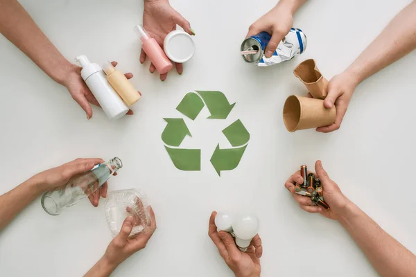 Vaya verde con el reciclaje. Vista superior de muchas manos sosteniendo diferentes residuos, tipos de basura con signo de reciclaje de papel en el centro sobre fondo blanco Imagen de stock