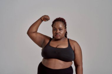 Vücudumu. Şişkin, ayrıca siyah spor giyim giymiş Afro-Amerikan kadın kameraya bakıyor, stüdyoda gri arka planda güçlü bir kol gösteriyor. Spor anlayışı, sağlıklı yaşam tarzı, vücut pozitifliği, eşitlik