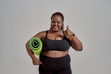 Her Güzel Vücut. Neşeli tombul, artı spor giyimli Afro-Amerikan kadın, yeşil yoga minderini gri arka planda tutarak yeşil yoga minderini tutuyor.