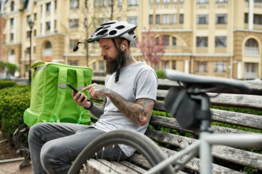 Just try it. Brutal bearded delivery man in helmet with thermo bag or backpack checking order using smartphone, sitting on the bench outdoors. Courier, delivery service concept clipart