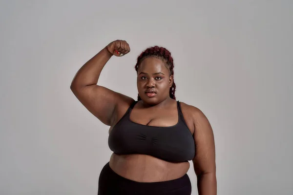 My body. Plump, plus size african american woman in black sportswear looking at camera, showing strong arm in studio over grey background. Concept of sport, healthy lifestyle, body positive, equality — Stock Photo, Image