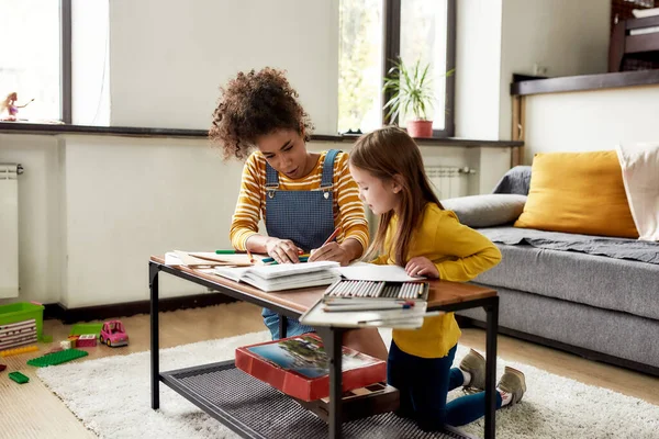 Dedicación. Niña caucásica pasando tiempo con niñera afroamericana. Están dibujando, aprendiendo a escribir cartas, sentados en el suelo —  Fotos de Stock