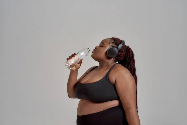 Vigyázz a testedre. Plump, plusz méretű afro-amerikai nő sportruházatban és fejhallgató ivóvíz az üvegből, pózol stúdióban szürke háttér — Stock Fotó