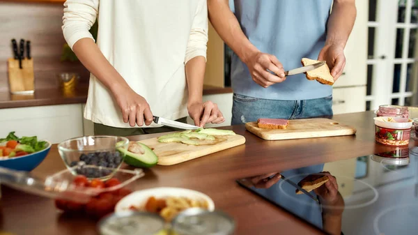 两个素食主义者一起在厨房里准备健康的饭菜。男人在烤面包上涂鹰嘴豆，女人在切鳄梨 — 图库照片