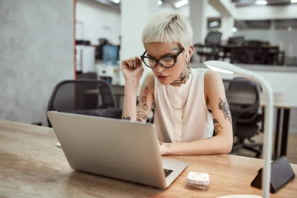 Пытаюсь найти загрязнение. Задумчивая молодая блондинка татуированная бизнесвумен в очках, глядя на экран ноутбука и думая о чем-то, сидя на своем рабочем месте — стоковое фото