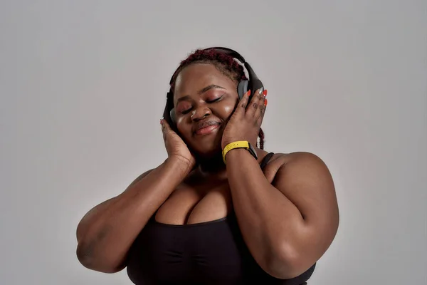 Musik. Plump, plus size afrikanisch amerikanische Frau in Sportbekleidung und Kopfhörer Musik hörend, im Studio vor grauem Hintergrund stehend. Konzept des Sports, gesunder Lebensstil, positiver Körper, Gleichberechtigung — Stockfoto