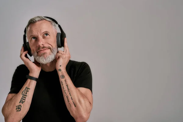 Sognavo. Uomo muscoloso di mezza età in t-shirt nera e cuffie alzando lo sguardo mentre ascolta musica, in posa in studio su sfondo grigio — Foto Stock