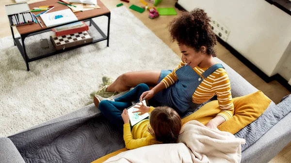Tecnologias para crianças. afro-americana baby sitter divertido caucasiano bonito menina. Kid está desenhando usando tablet pc, sentado no sofá — Fotografia de Stock