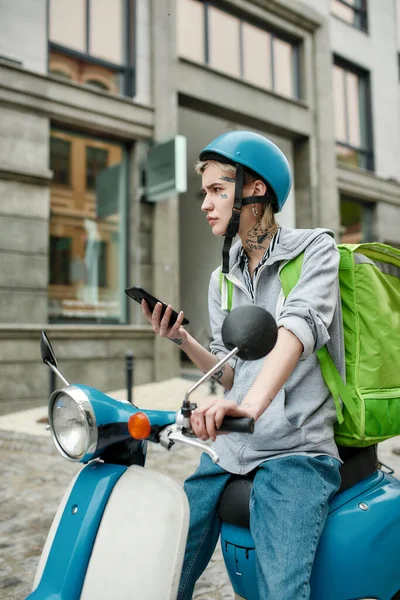 年轻女子头戴头盔，背着热身背包，一边用智能手机一边用摩托车运送食物 — 图库照片