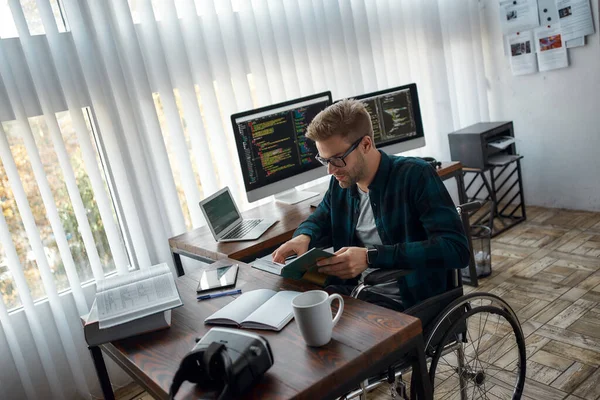 Me estoy desarrollando. Joven desarrollador web masculino en silla de ruedas leyendo un libro mientras está sentado en su lugar de trabajo con múltiples pantallas de computadora en el fondo — Foto de Stock