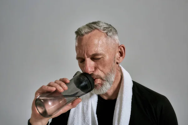 Uomo muscoloso di mezza età in t-shirt nera che chiude gli occhi mentre beve acqua dalla bottiglia, posando in studio su sfondo grigio — Foto Stock