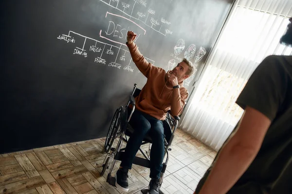 Brainstormen. Jonge professionele ontwerper in een rolstoel bespreekt iets met zijn collega terwijl hij naast het schoolbord zit met presentatie in het moderne kantoor — Stockfoto