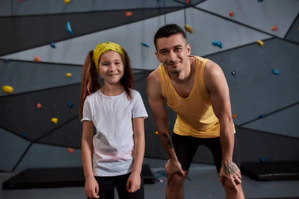 Genç erkek eğitmen ve aktif küçük kız kameraya bakıyor, yapay tırmanma duvarına karşı duruyor. Spor hayatı ve kaya tırmanışı kavramı — Stok fotoğraf