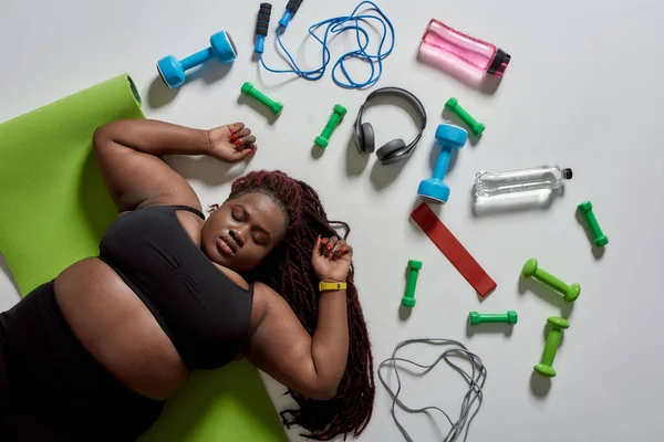 Mejor cambio. Vista superior de la gorda cansada, más tamaño mujer afroamericana en ropa deportiva acostada en la esterilla de yoga después del entrenamiento con deportes, equipo de fitness y auriculares cerca de ella sobre fondo gris — Foto de Stock