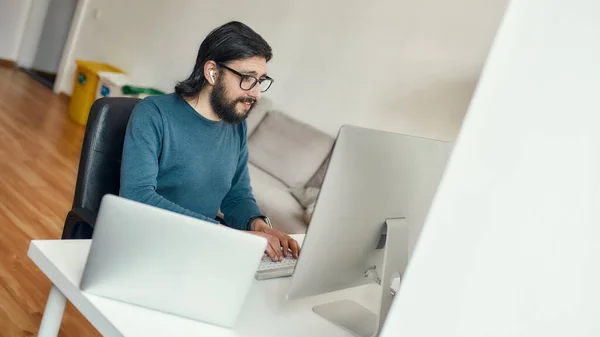 Jeune homme barbu concentré caucasien assis à son lieu de travail et travaillant avec l'ordinateur à distance, travaillant de la maison. Freelance, bureau à domicile — Photo