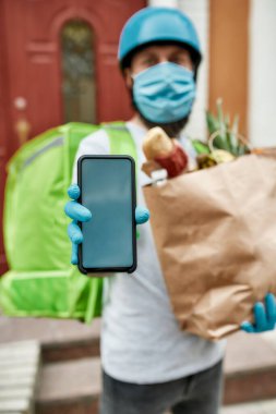 Sakallı teslimatçı, Covid19 virüsünün ortaya çıkmasından dolayı maske ve eldiven takıyor. Akıllı telefon gösteriyor.