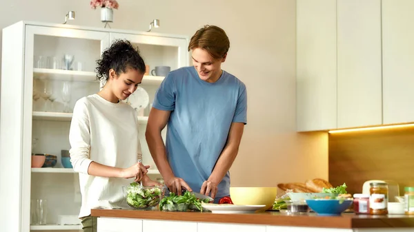 Hombre joven cortando verduras mientras la mujer lanza una ensalada. Vegetarianos preparando comida saludable en la cocina juntos. Vegetarianismo, concepto de comida saludable — Foto de Stock