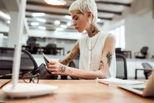 Looking for sales report. Young blonde tattooed businesswoman with short haircut trying to find something in her handbag while sitting at her desk in office