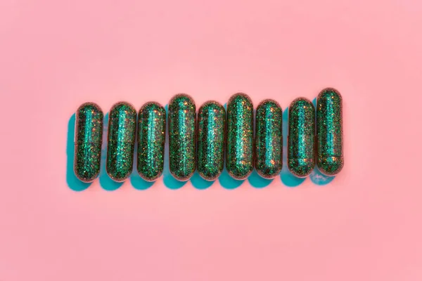 Concetto creativo con molte pillole glitter verdi distese in fila orizzontalmente isolate su sfondo rosa pastello. Stile minimale, concetto di arte — Foto Stock