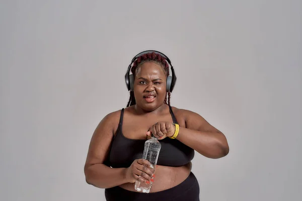 Plump, plus size afrikanische Amerikanerin in Sportbekleidung und Kopfhörer lächelnd, Flasche Wasser öffnend, posierend im Studio vor grauem Hintergrund — Stockfoto