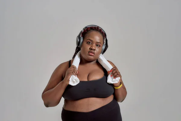 Plump, plus size afrikanisch-amerikanische Frau in Sportbekleidung und Kopfhörer mit Handtuch um den Hals posiert im Studio vor grauem Hintergrund — Stockfoto