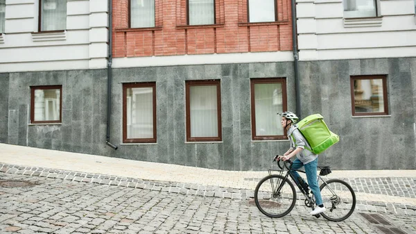 Junge Zustellerin mit Helm, Thermosack oder Rucksack, die mit dem Fahrrad durch die Stadt fährt und Essen ausliefert. Kurier, Lieferservice-Konzept — Stockfoto