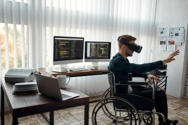 Zijaanzicht van een jonge mannelijke webontwikkelaar in een rolstoel met een virtual reality bril terwijl hij op zijn werkplek in een modern kantoor zit. Meerdere computerschermen met programmacode — Stockfoto