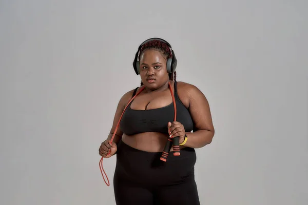 Plump, plus size afrikanisch amerikanische Frau in Sportbekleidung und Kopfhörer mit Springseil, posiert im Studio vor grauem Hintergrund. Konzept des Sports, gesunder Lebensstil, positiver Körper, Gleichberechtigung — Stockfoto