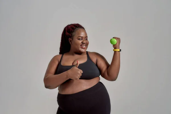 Plump, plus size afrikanisch amerikanische Frau in Sportbekleidung zeigt Daumen nach oben während des Trainings, hält grüne Hantel im Studio über grauem Hintergrund — Stockfoto