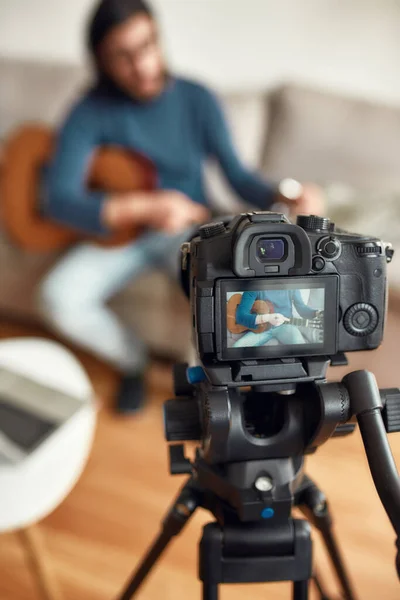Guitar lessons. Young man sitting on sofa at home and teaching how to play guitar. Recording webinar at home. Learning music online. Focus on a camera — Stock Photo, Image