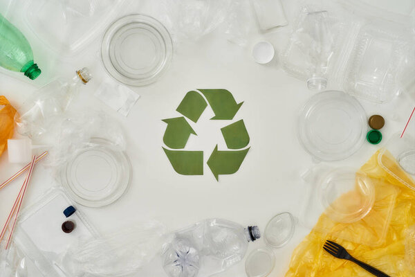 Flatlay composition with different empty plastic bottles, containers and recycling sign made of paper in the center over white background