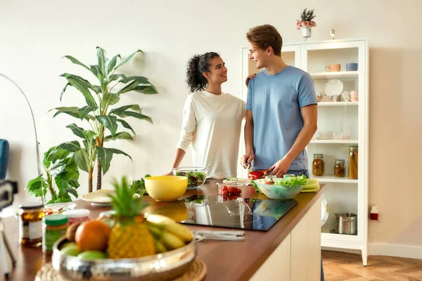 Νεαρός κοιτάζει την κοπέλα του ενώ κόβει λαχανικά. Χορτοφάγοι ετοιμάζουν υγιεινό γεύμα στην κουζίνα μαζί. Χορτοφαγία, υγιεινή διατροφή έννοια — Φωτογραφία Αρχείου