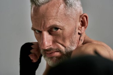 Orta yaşlı atletik bir adam, boks yaparken agresif görünen bir boksör, gri arka plan üzerinden atölyede eğitim görüyor. Muay Thai, Boks veya Kickboks konsepti