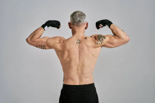 Mezza nuda uomo atletico di mezza età, kickboxer mostrando il suo corpo, durante l'esercizio, praticare pugni in studio su sfondo grigio — Foto Stock
