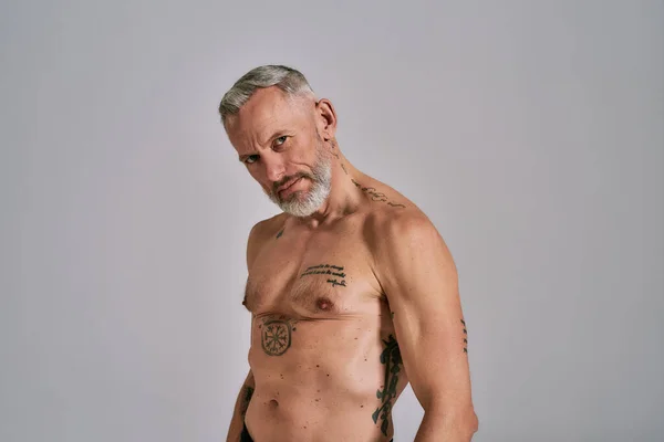 Félmeztelen középkorú izmos férfi néz kamera, mutatja a testét, miközben pózol a stúdióban szürke háttér — Stock Fotó