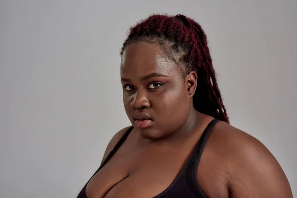 Nahaufnahme einer dicken, plus size afrikanisch-amerikanischen Frau in schwarzer Sportbekleidung, die in die Kamera schaut, während sie im Studio vor grauem Hintergrund posiert — Stockfoto