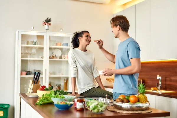Pareja feliz, vegetarianos preparando comida saludable, sandwhich, ensalada en la cocina juntos. Chico alimentando a su novia frutos secos — Foto de Stock