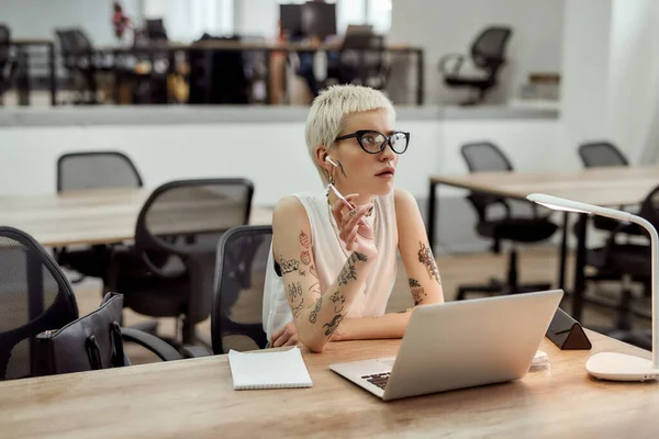 Az üzleten gondolkodom. Portré fiatal szőke tetovált üzletasszonyról szemüvegben, rövid hajjal, vezeték nélküli fülhallgatóval és gondolkodás közben a modern irodában. — Stock Fotó