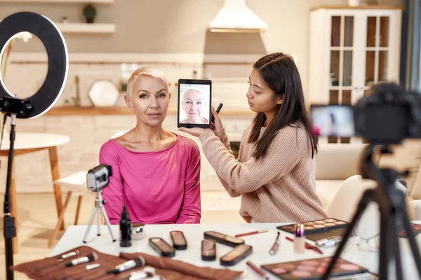 Jól néz ki. Fiatal női blogger, sminkes segítségével tabletta pc, tartja előtt középkorú nő arca alkalmazása után smink — Stock Fotó