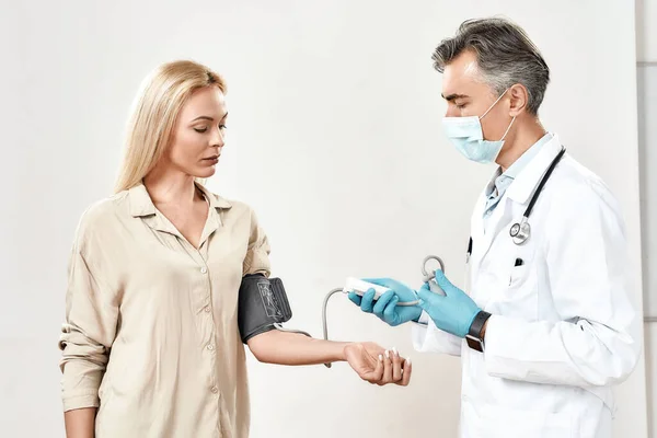 健康診断だ。若い女性の血圧を測定保護顔マスクと青滅菌手袋を身に着けている男性成熟した医師,彼の女性患者 — ストック写真