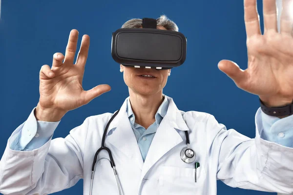 Foto de cerca del médico masculino en uniforme médico usando gafas de realidad virtual mientras está de pie contra el fondo azul marino —  Fotos de Stock