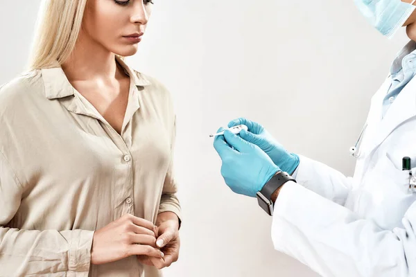 Ziyaret kliniği. Mavi steril eldivenli profesyonel erkek doktor ve elinde elektronik termometre tutan koruyucu maske genç bayan hastanın ısısını ölçüyor. — Stok fotoğraf