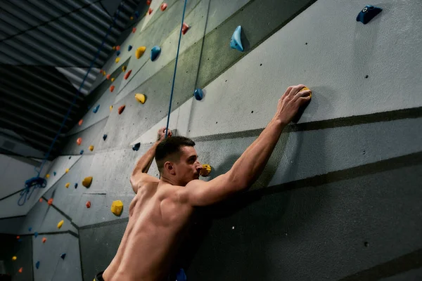 Homem atlético jovem olhando focado, enquanto o treinamento na parede de escalada artificial dentro de casa — Fotografia de Stock