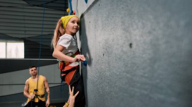 Erkek eğitmen öğretmenlik yapıyor, yapay tırmanma duvarı eğitimi alırken güvenlik ekipmanı ve koşum takımı takan bir kıza bakıyor.