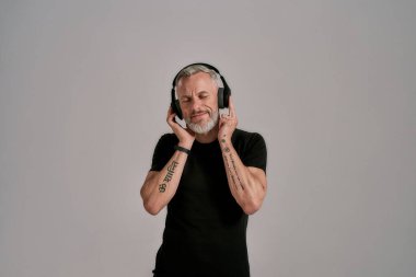 Orta yaşlı, siyah tişörtlü ve kulaklıklı bir adam stüdyoda gri arka planda müzik dinlerken gözlerini kapatıyor.