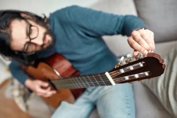 Primo piano del giovane caucasico seduto sul divano a casa e che regola la chitarra acustica. Focus principale su una chitarra. Suonare strumento musicale. — Foto Stock