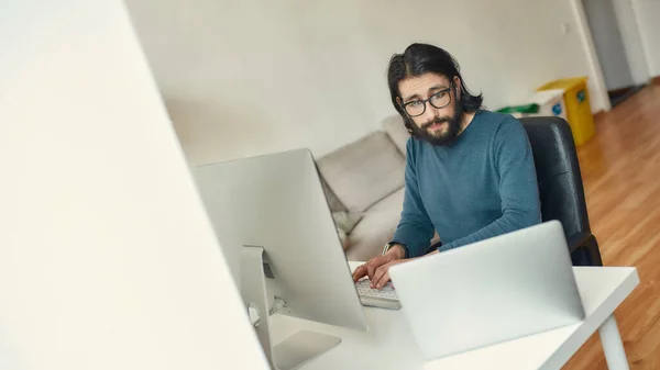 Multitasking. Ung kaukasisk koncentrerad skäggig man sitter på sin arbetsplats och arbetar på distans, med hjälp av stationär dator och bärbar dator — Stockfoto