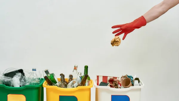 Hand in red glove throwing paper into one of three colorful green, yellow and white recycle garbage bins full of rubbish isolated on white background. Waste separation concept — Stock Photo, Image